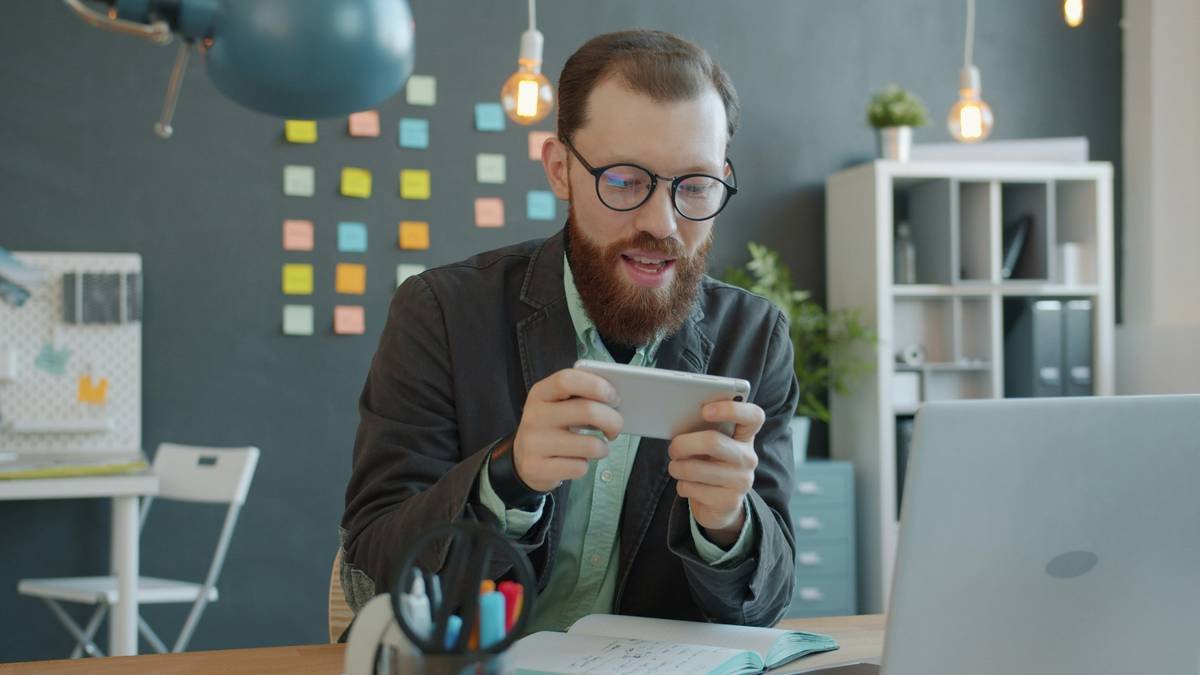 Joyful guy is enjoying mobile video game playing alone in creative office relaxing during break. Smartphone applications, people and relaxation concept.